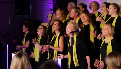 Ein Teil des Albertinen Mitarbeitenden Gospelchors singt aus voller Kehle in festlicher Kleidung mit grünen Schals im festlich beleuchteten Kirchenraum der Kirche am Albertinen Krankenhaus in Hamburg.