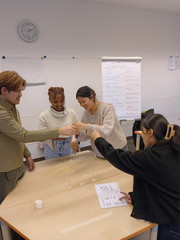 Vier Auszubildende koordinieren Handgriffe beim Bau eines Spaghetti-Turms; im Hintergrund steht ein Flipchart zum Thema Teamarbeit.