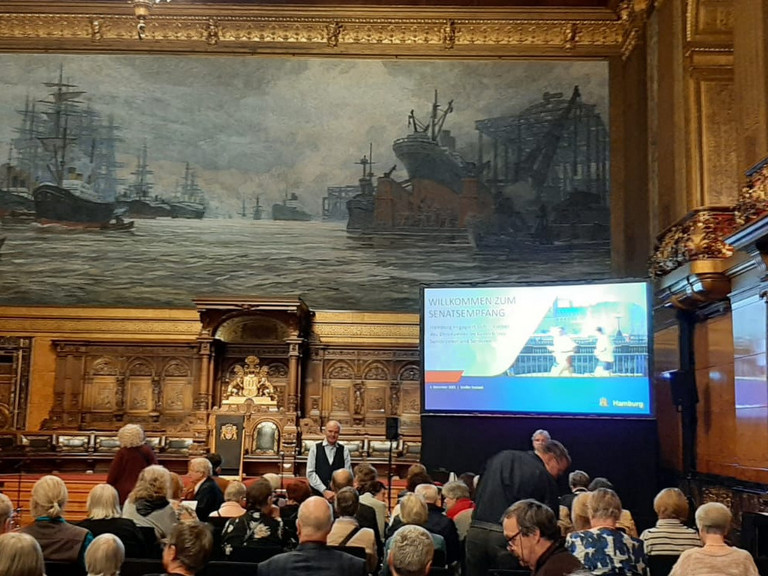 Blick über die Stuhlreihen mit Publikum im Festsaal des Hamburger Rathauses Richtung Podium mit einem großen Monitor zum Senatsempfang für ehrenamtlich engagierte Menschen mit Ehrung der Ehrenamtlichen im Albertinen Haus Hamburg