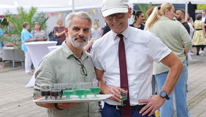 Pflegedirektor Alberto da Silva Correia steht mit einem Serviertablett mit eingefärbten Cocktails neben Ralf Zastrau beim Albertinen-Sommerfest für die Mitarbeitenden aus den unterschiedlichen Einrichtungen der Immanuel Albertinen Diakonie am Standort Schnelsen auf dem Platz vor der Kirche am Albertinen Krankenhaus.