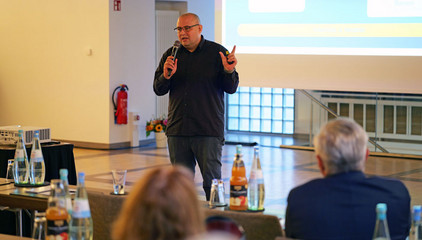 Dr. Schöps spricht mit einem Mikrofon in der Hand neben einer Projektionsleinwand zum Publikum bei einem Vortrag beim Einführungssymposium „Die Zukunft ist alt!“ von Priv.-Doz. Dr. Gödel, Chefarzt der Klinik für Geriatrie im Albertinen Zentrum für Altersmedizin im Albertinen Krankenhaus in Hamburg. Dr. Schöps spricht mit einem Mikrofon in der Hand neben einer Projektionsleinwand zum Publikum bei einem Vortrag beim Einführungssymposium „Die Zukunft ist alt!“ von Priv.-Doz. Dr. Gödel, Chefarzt der Klinik für Geriatrie im Albertinen Zentrum für Altersmedizin im Albertinen Krankenhaus in Hamburg.