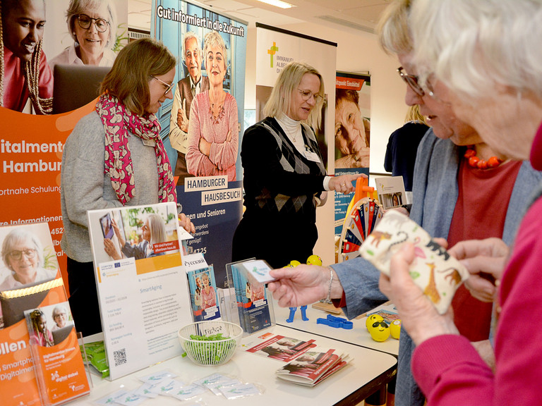 Zwei Mitarbeitende stehen vor Informationsplakaten am Infostand Albertinen Haus auf dem Eimsbütteler Seniorentag, während zwei Frauen die ausgelegten Informationsmaterialien und Giveaways auf dem Tisch näher betrachten