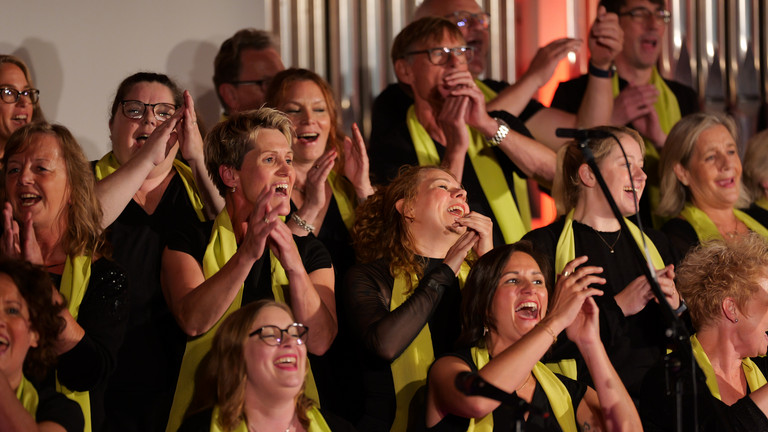 Mehrere in die Hände klatschende Sängerinnen und Sänger des Albertinen Mitarbeitenden Gospelchors im bunt beleuchteten Kirchenraum der Kirche am Albertinen Krankenhaus in Hamburg. 