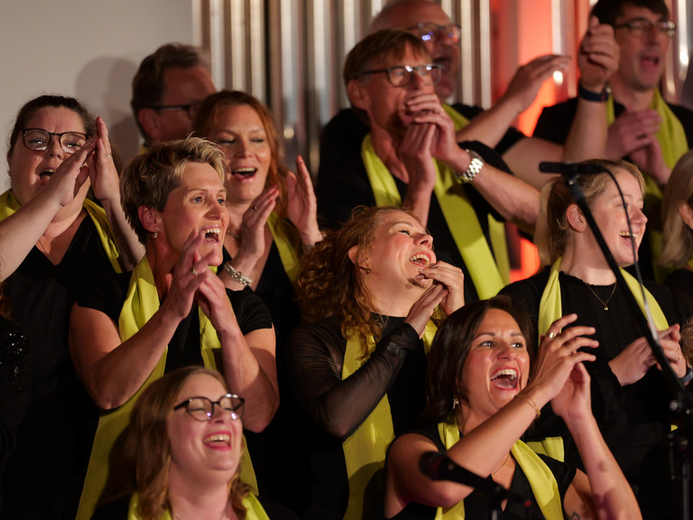 Mehrere in die Hände klatschende Sängerinnen und Sänger des Albertinen Mitarbeitenden Gospelchors im bunt beleuchteten Kirchenraum der Kirche am Albertinen Krankenhaus in Hamburg. 