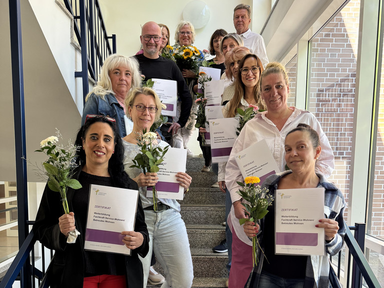 Gruppenfoto 11. Jahrgang der beruflichen Weiterbildung Fachkraft Service-Wohnen / Betreutes Wohnen mit Abschlusszertifikaten und Blumensträußen auf dem Treppenaufgang