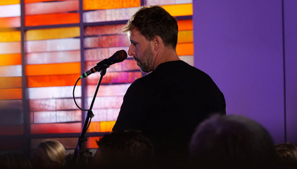 Chorleiter Sören Schröder des Albertinen Mitarbeitenden Gospelchors steht am Mikrfon vor einem bunten Kirchenfenster im festlich beleuchteten Kirchenraum der Kirche am Albertinen Krankenhaus in Hamburg.