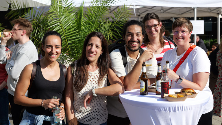 Mitarbeitende an einem weiß gedeckten Stehtisch mit Getränken und Burgern blicken lachend in die Kamera beim Albertinen-Sommerfest für die Mitarbeitenden aus den unterschiedlichen Einrichtungen der Immanuel Albertinen Diakonie am Standort Schnelsen auf dem Platz vor der Kirche am Albertinen Krankenhaus.