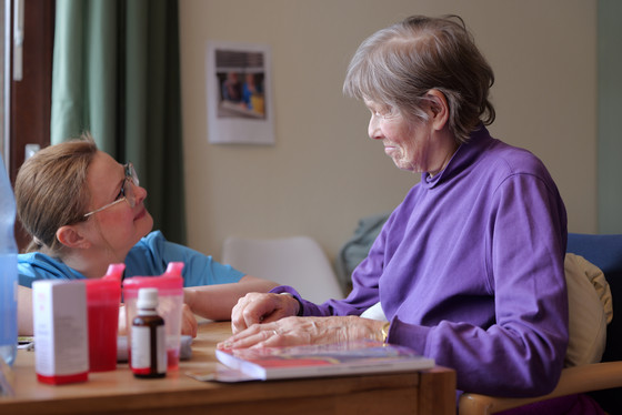 Pflegefachkraft Inna Gaberkorn unterhält sich am Tisch kniend mit einer am Tisch sitzenden Bewohnerin nach dem gemeinsam erfolgreich bewältigten Transfer vom Rollstuhl in den Lehnstuhl im Präventionspflegeprojekt „SGB Reha“ im Albertinen Haus in Hamburg